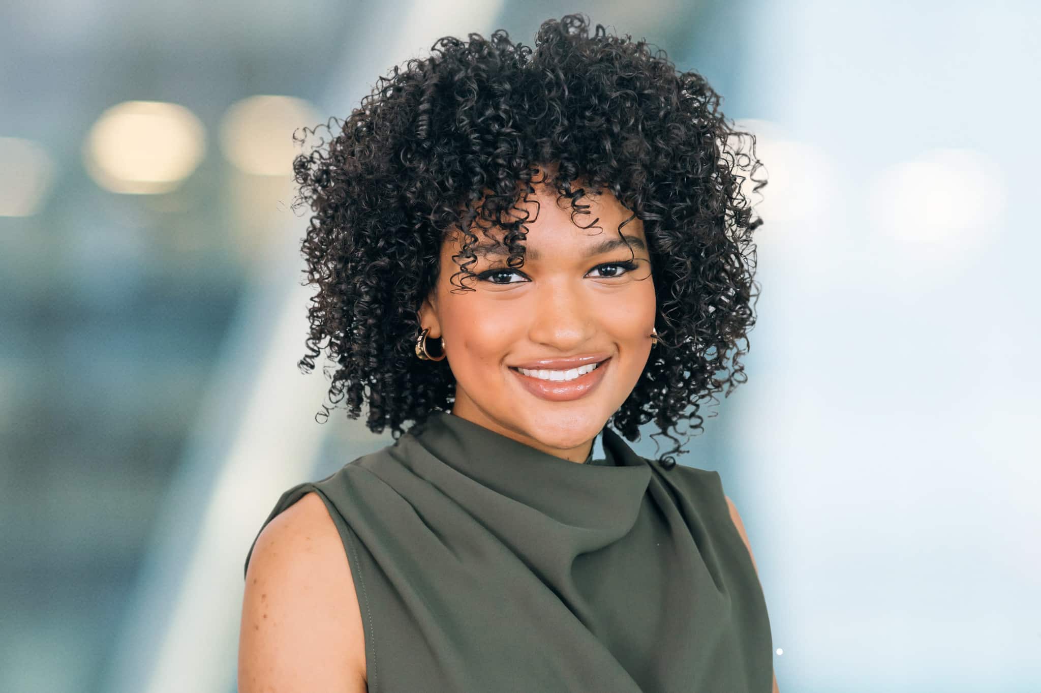 Marah Burns A person with curly hair smiles at the camera, wearing a sleeveless green top. The background is softly blurred with bright lights.