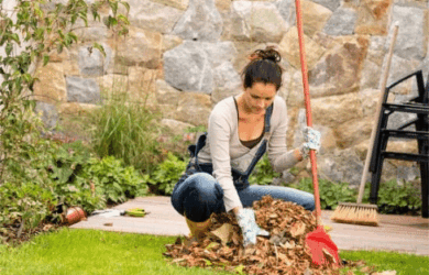 A woman wearing gloves and casual clothes is raking a pile of autumn leaves on a wooden deck in a garden. She holds a red rake and is surrounded by green plants. In the background, stacked chairs and a stone wall are visible.