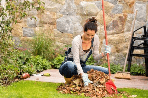 A woman wearing gloves and casual clothes is raking a pile of autumn leaves on a wooden deck in a garden. She holds a red rake and is surrounded by green plants. In the background, stacked chairs and a stone wall are visible.