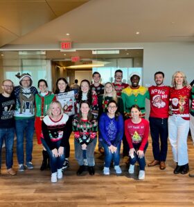 A group of 16 people stands together in a brightly lit office space, all wearing festive holiday sweaters. They are smiling, and some are posing with playful gestures. The room features modern decor and large windows.