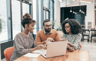 Three people sit around a wooden table, engaged in discussion, looking at a laptop. Two women with curly hair are on either side of a man with glasses and a beard. The setting is a modern office with large windows.