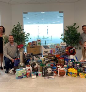 Five people pose in an office setting with a collection of toys, games, and sporting goods displayed on the floor. The group is smiling, and there are plants and office furniture in the background.