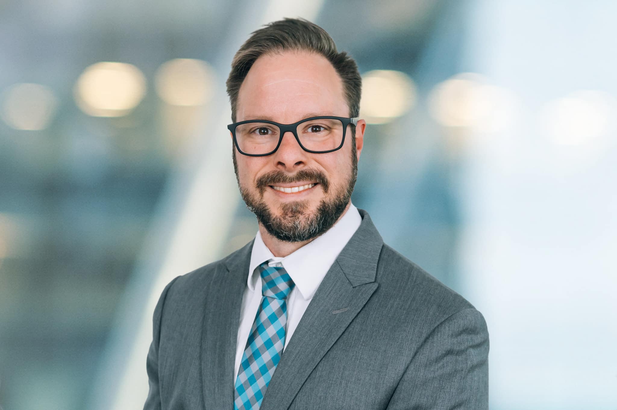 Man with short hair, beard, and glasses, wearing a gray suit, white shirt, and blue checkered tie, smiling against a blurred indoor background.