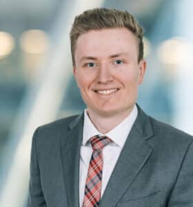 A person in a suit and red plaid tie smiles at the camera. The background is softly blurred and features lights, giving the image a professional appearance.