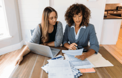 Two women sitting at a table reviewing financial documents. One woman has a laptop open, while the other holds a paper and a calculator. Several sheets of paper and envelopes are spread across the table in a bright room.
