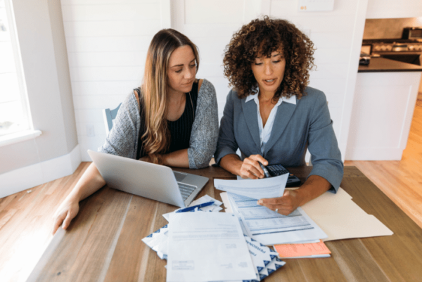 Two women sitting at a table reviewing financial documents. One woman has a laptop open, while the other holds a paper and a calculator. Several sheets of paper and envelopes are spread across the table in a bright room.