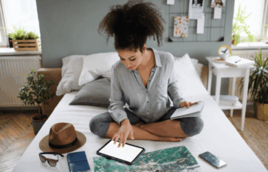 A woman with curly hair sits cross-legged on a bed, using a tablet. She holds a notepad and is surrounded by travel items: a map, passport, hat, smartphone, and camera. The room is bright with potted plants and a bulletin board in the background.