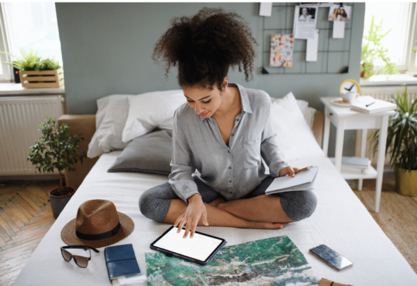 A woman with curly hair sits cross-legged on a bed, using a tablet. She holds a notepad and is surrounded by travel items: a map, passport, hat, smartphone, and camera. The room is bright with potted plants and a bulletin board in the background.