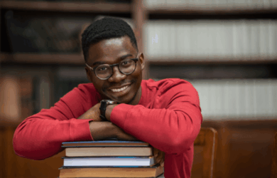 A person wearing glasses and a red sweater is smiling and leaning on a stack of books in a library setting. Shelves filled with books can be seen in the background.