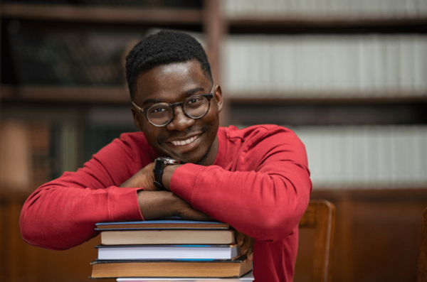 A person wearing glasses and a red sweater is smiling and leaning on a stack of books in a library setting. Shelves filled with books can be seen in the background.