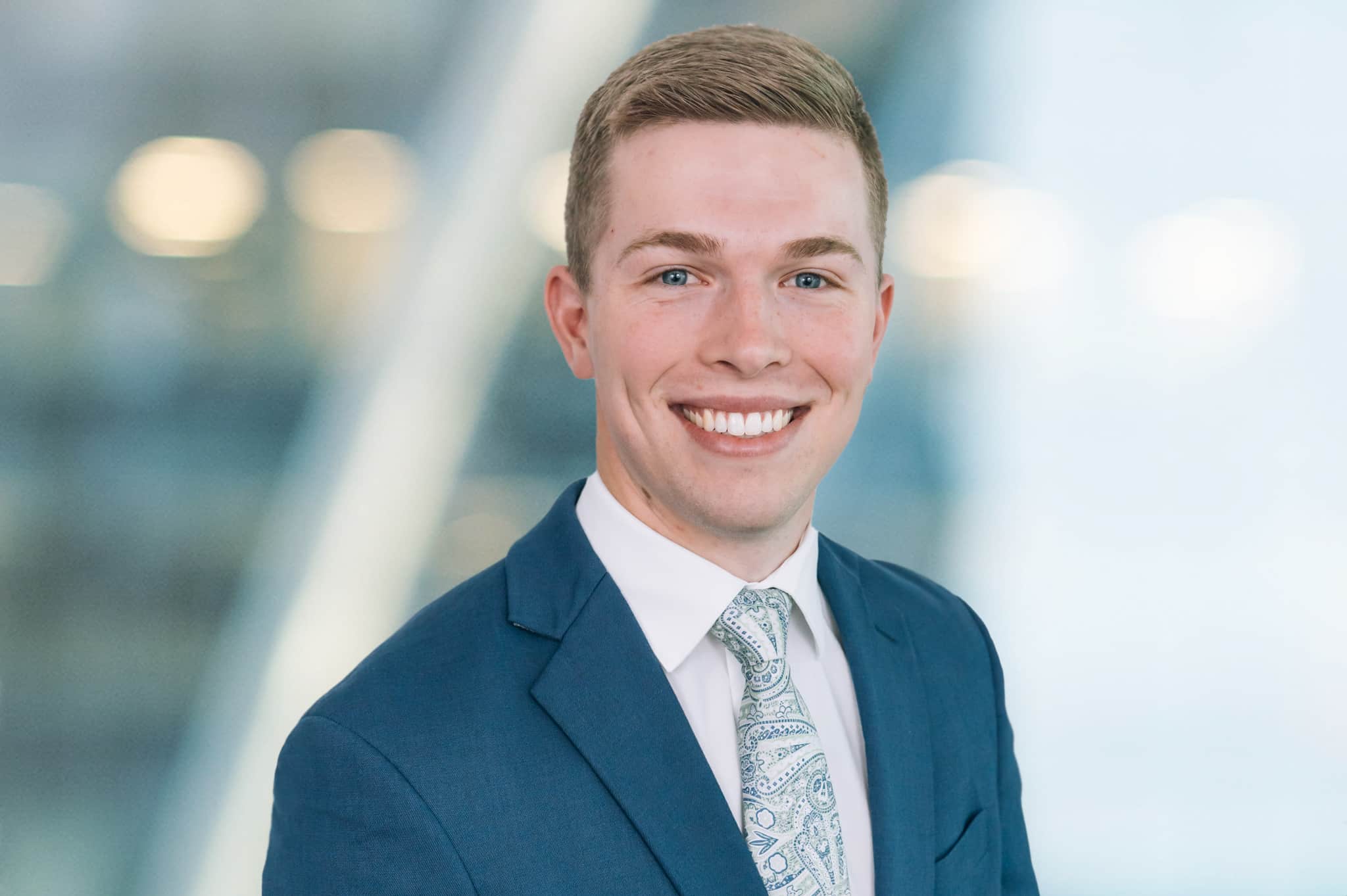 A person in a blue suit and patterned tie smiles warmly. The background is softly blurred with bokeh lights.