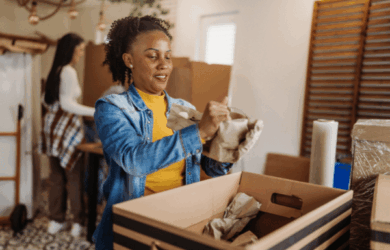 A woman in a denim jacket and yellow shirt is wrapping items in paper and packing them into a large box. Another person is in the background. The setting appears to be a home with more boxes visible, suggesting they are preparing to move.