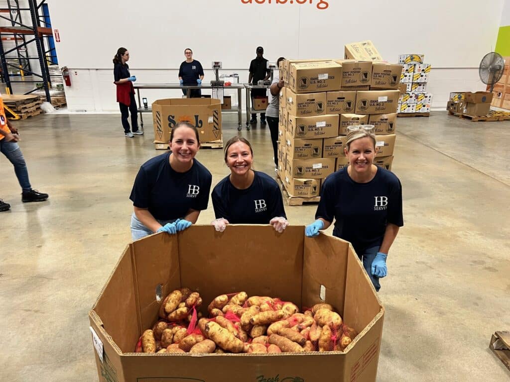 Three people in navy shirts and blue gloves smile while kneeling beside a large box of sweet potatoes in a warehouse. Other people work in the background, surrounded by boxes and supplies.
