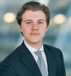 A young man in a suit and tie stands against a blurred background. He has short brown hair and is looking directly at the camera with a subtle smile.