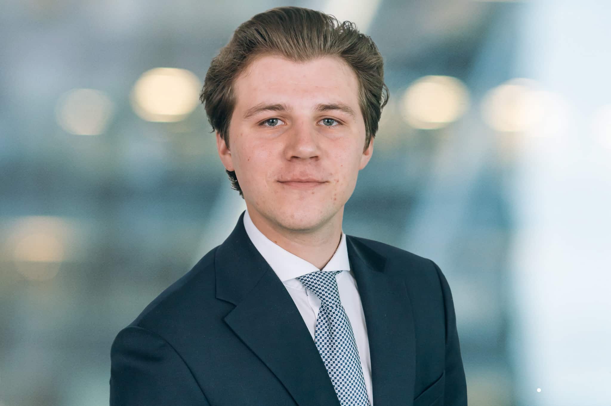 A young man in a suit and tie stands against a blurred background. He has short brown hair and is looking directly at the camera with a subtle smile.