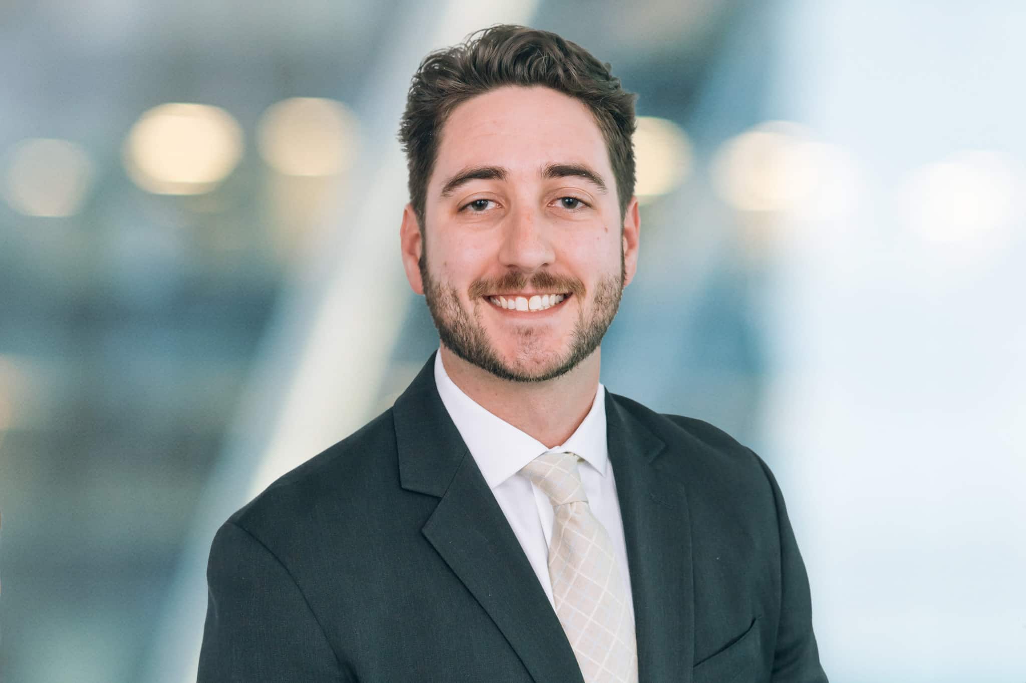 A man with short dark hair and a beard is smiling. He is wearing a white shirt, a light-colored tie, and a dark suit jacket. The background is blurred, featuring soft lights and a modern setting.