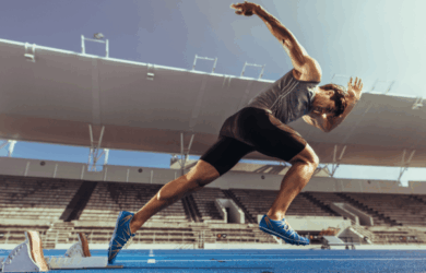 A sprinter launches off the starting blocks on a blue track in an empty stadium. The athlete is wearing a gray tank top, black shorts, and blue running shoes, captured in mid-stride with dramatic sunlight highlighting the scene.