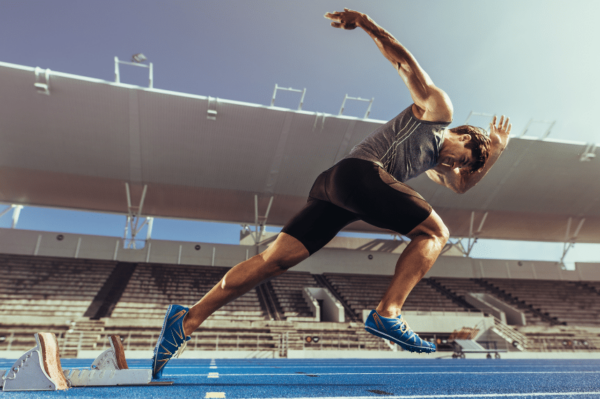 A sprinter launches off the starting blocks on a blue track in an empty stadium. The athlete is wearing a gray tank top, black shorts, and blue running shoes, captured in mid-stride with dramatic sunlight highlighting the scene.