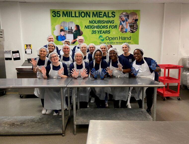 A group of people wearing aprons and hairnets, smiling and posing with gloved hands raised in a kitchen setting. Behind them is a banner celebrating 35 Million Meals from Open Hand, highlighting 35 years of service.