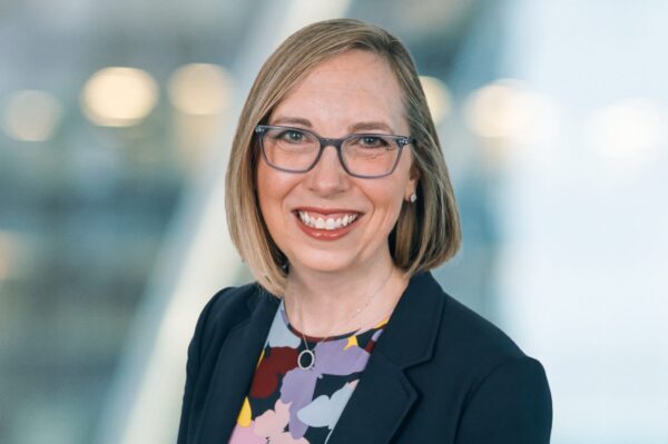 A smiling person with shoulder-length blonde hair and glasses is wearing a dark blazer over a colorful patterned top. The background is softly blurred with light and shadow contrasts.