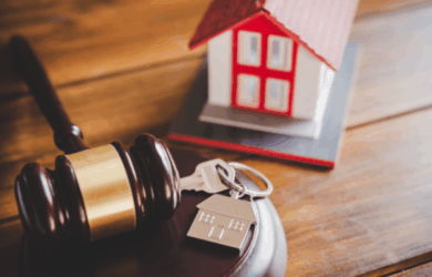 A wooden gavel rests beside a set of house keys with a house-shaped keychain. In the background, there is a small model of a house with a red roof, symbolizing real estate or property law.