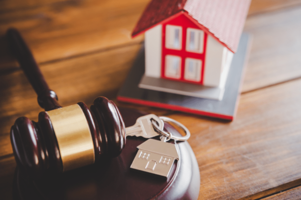 A wooden gavel rests beside a set of house keys with a house-shaped keychain. In the background, there is a small model of a house with a red roof, symbolizing real estate or property law.
