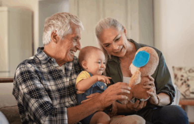 Older couple smiling and sitting with a baby on the sofa. The baby is playing with a stuffed toy. All appear joyful and engaged.