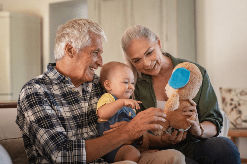 Older couple smiling and sitting with a baby on the sofa. The baby is playing with a stuffed toy. All appear joyful and engaged.
