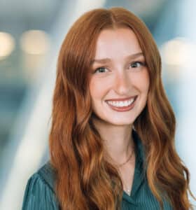 A person with long red hair and a green top smiles against a blurred background.