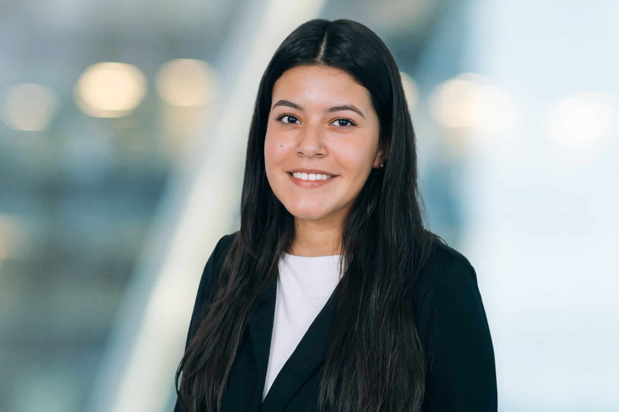 A woman with long dark hair is smiling, wearing a black blazer over a white top. The background is blurred with lights, giving a professional and airy atmosphere.
