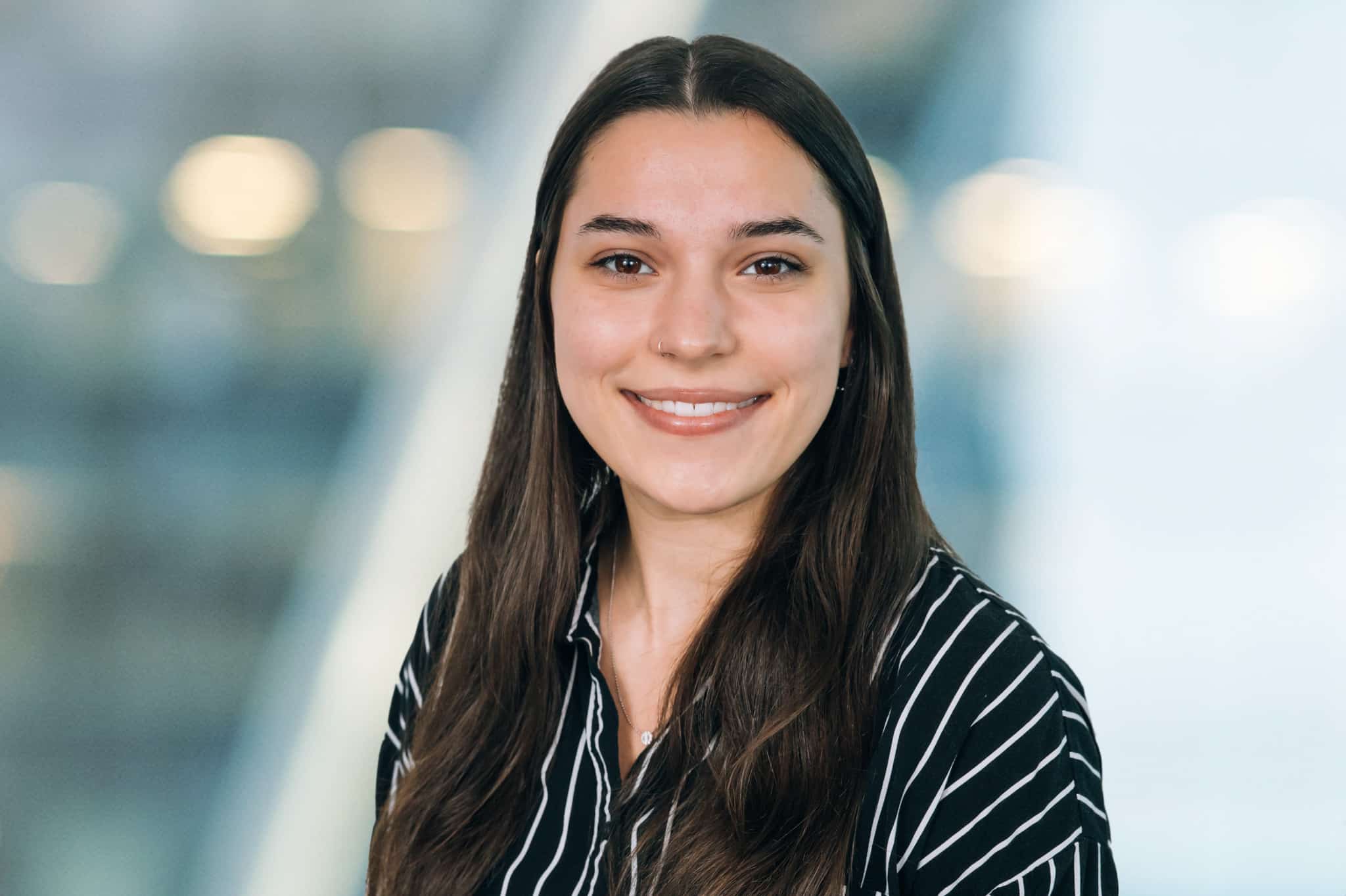 A person with long brown hair is smiling, wearing a black and white striped blouse. The background is softly blurred with some round lights visible, creating a professional yet approachable atmosphere.