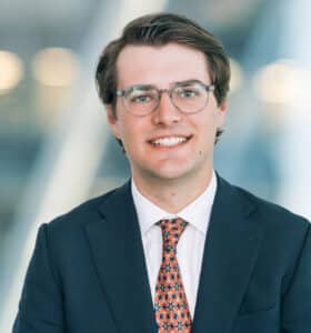 A person with brown hair and glasses, wearing a dark suit, a white shirt, and a patterned tie, smiles at the camera. The background is blurred, with abstract light shapes.
