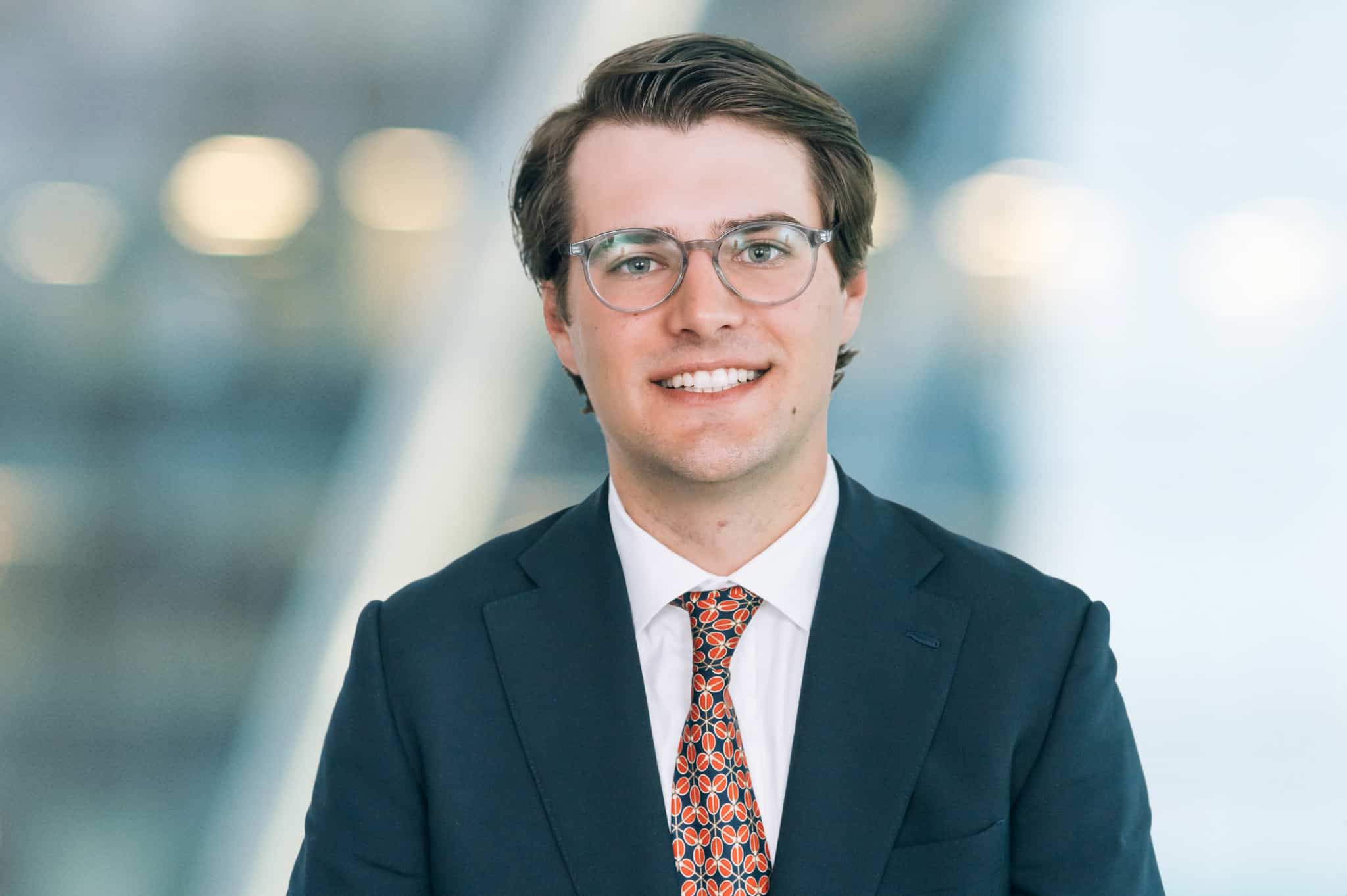 A person with brown hair and glasses, wearing a dark suit, a white shirt, and a patterned tie, smiles at the camera. The background is blurred, with abstract light shapes.
