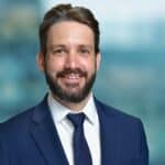 A man in a blue suit and tie smiles against a blurred blue and green background.