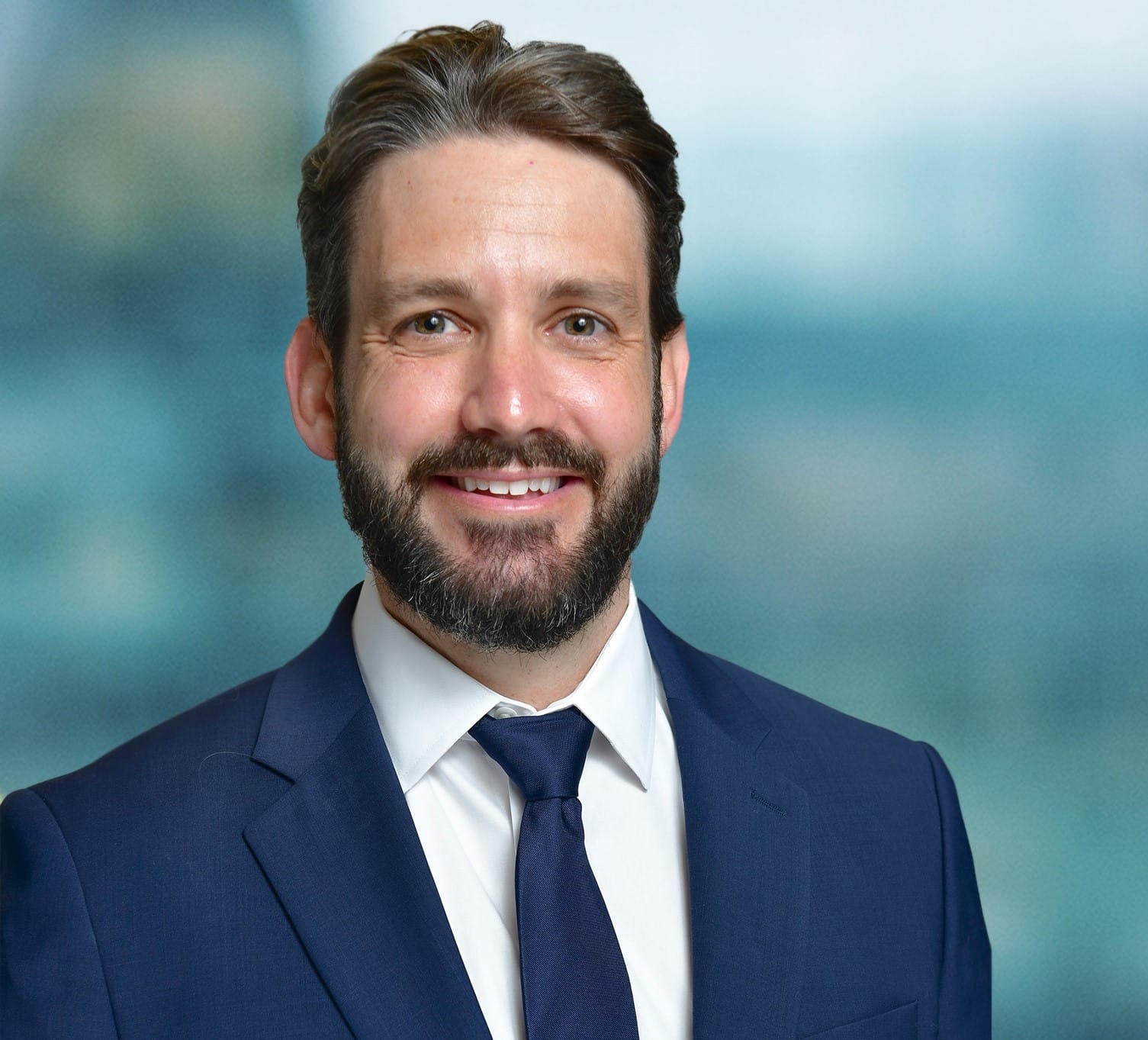 A man in a blue suit and tie smiles against a blurred blue and green background.