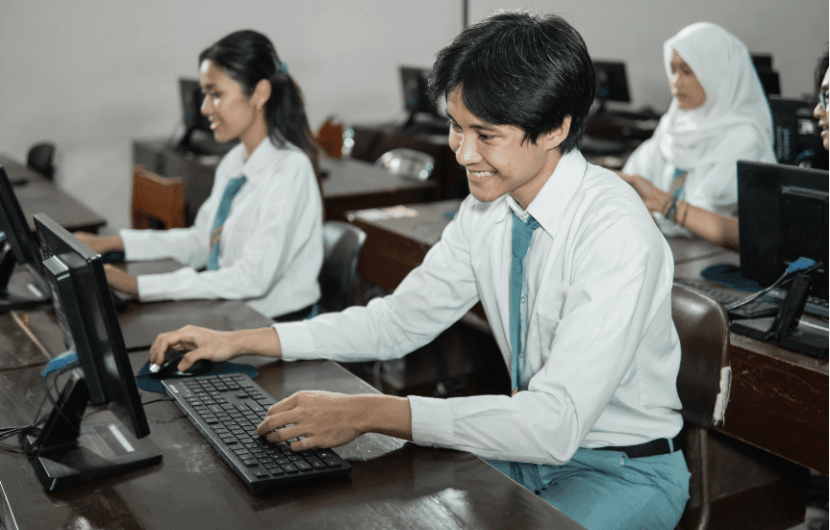 Students in uniform sit at computers in a classroom setting, appearing focused and engaged. A male student in the foreground smiles while typing. Several other students are visible in the background working at their stations.