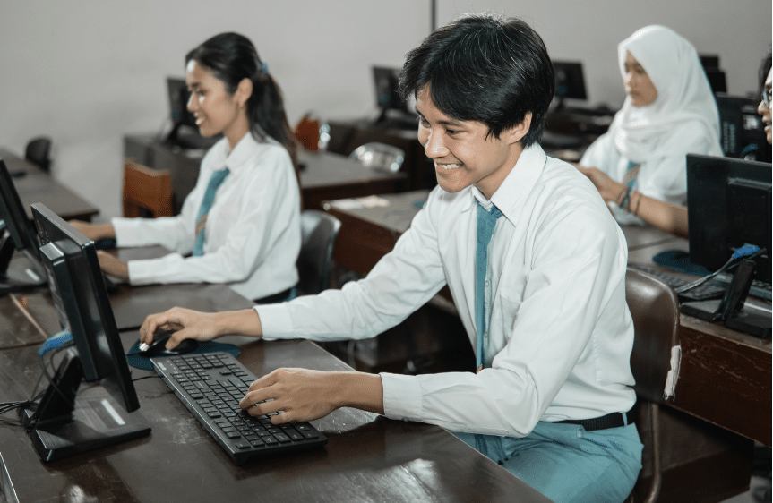 Students in uniform sit at computers in a classroom setting, appearing focused and engaged. A male student in the foreground smiles while typing. Several other students are visible in the background working at their stations.