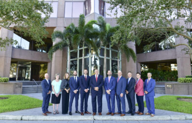 A group of twelve people, dressed in business attire, stand in front of a modern office building. Palm trees flank the entrance, and the building features large windows. The setting is daytime, with greenery visible around the area.