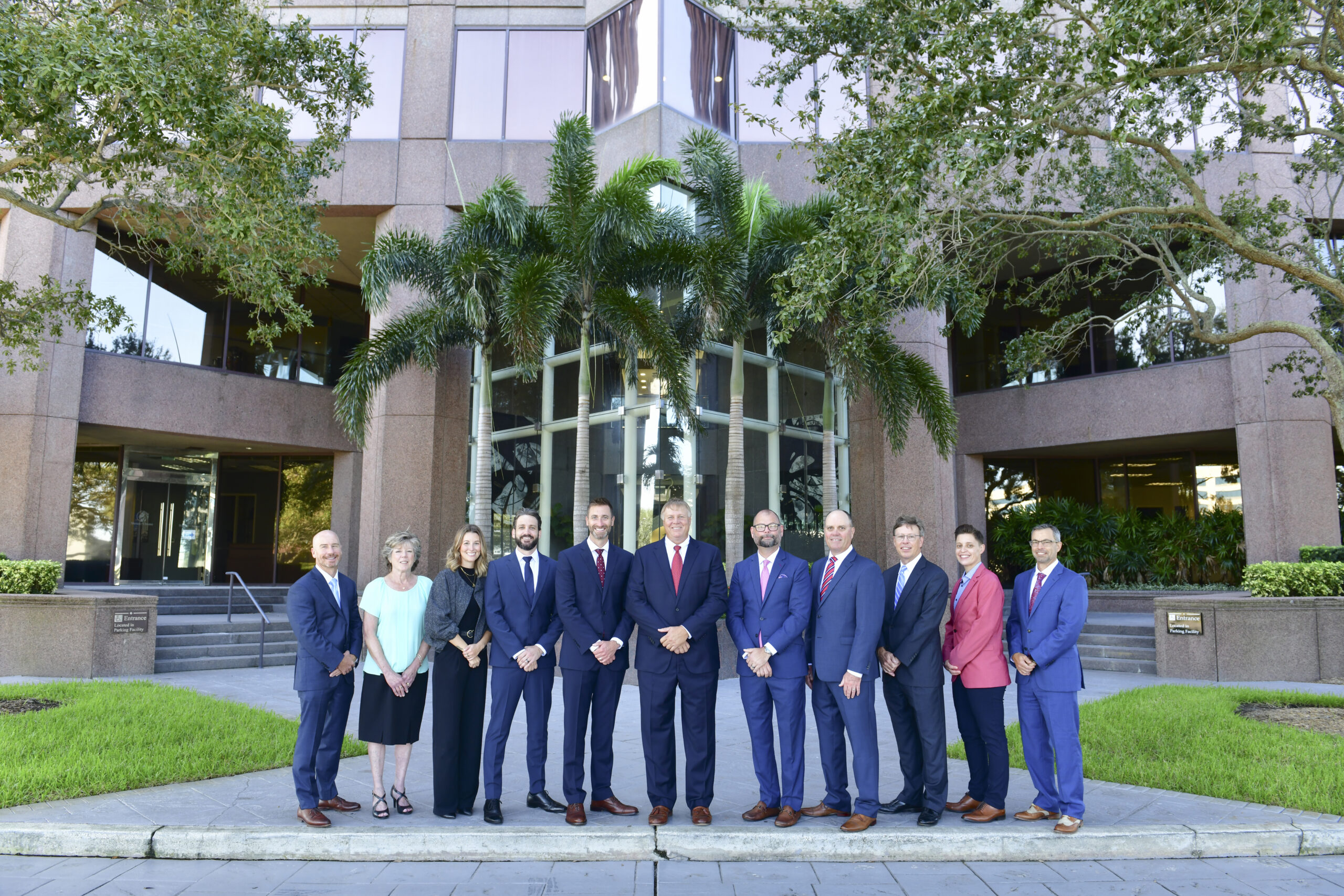 A group of twelve people, dressed in business attire, stand in front of a modern office building. Palm trees flank the entrance, and the building features large windows. The setting is daytime, with greenery visible around the area.