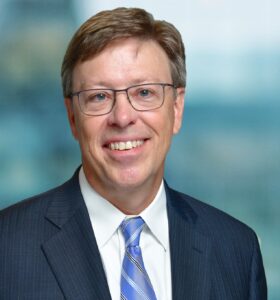 A person wearing glasses, a dark suit, blue and white striped tie, and a light blue shirt smiles in front of a blurred background.