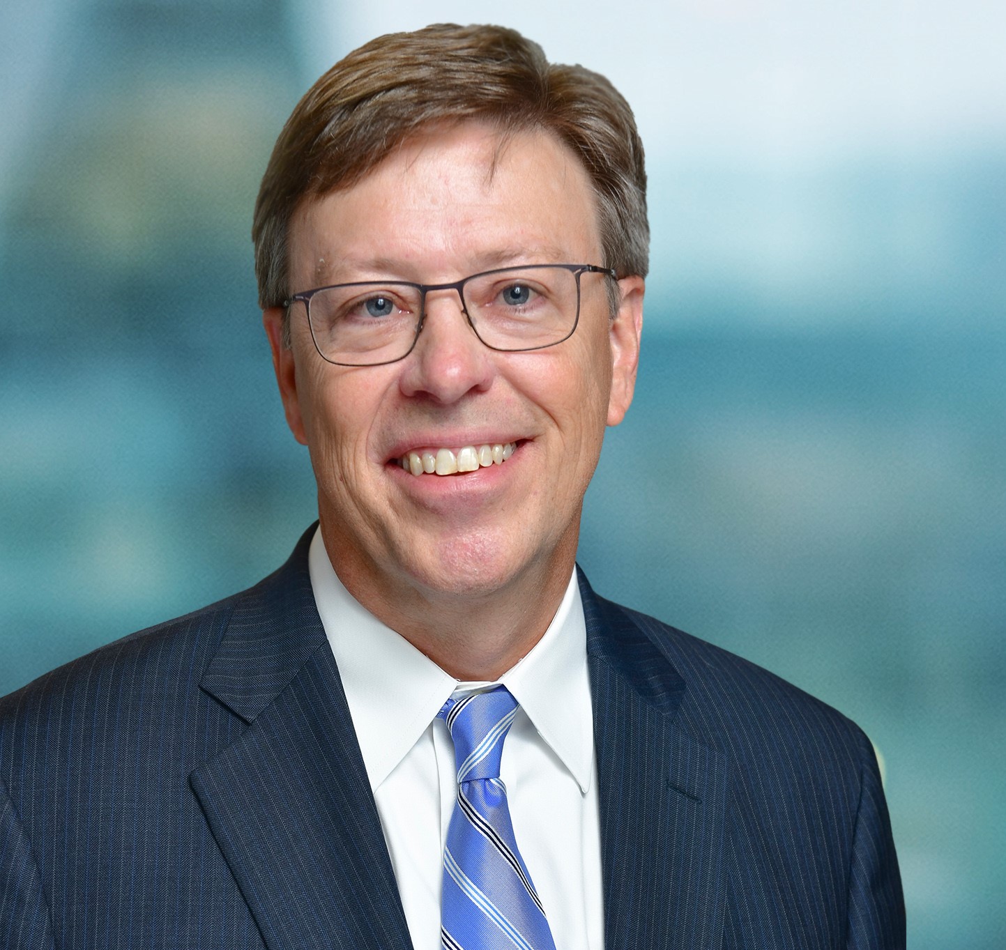 A person wearing glasses, a dark suit, blue and white striped tie, and a light blue shirt smiles in front of a blurred background.