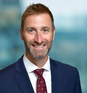 A man with short hair, a beard, and a friendly expression is wearing a navy suit, white shirt, and red patterned tie. The background is blurred, providing a soft, professional appearance.