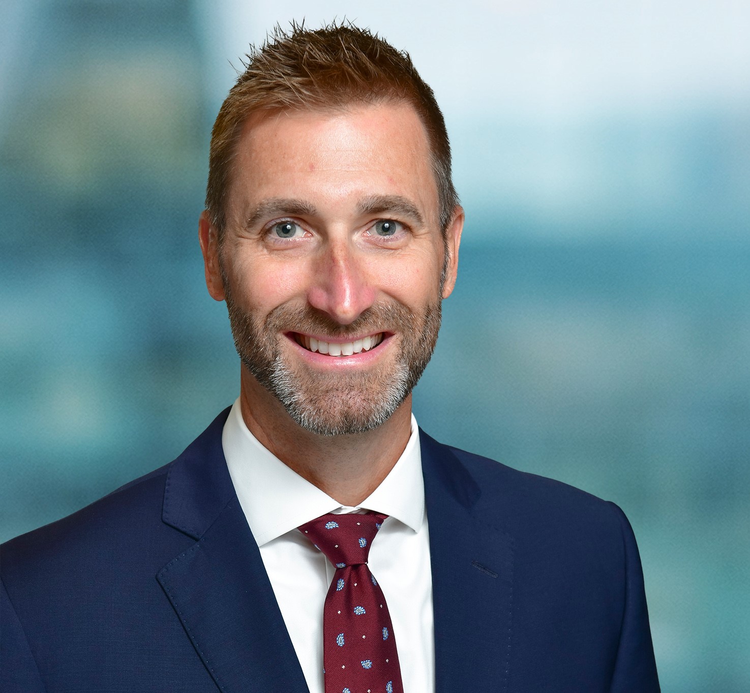 A man with short hair, a beard, and a friendly expression is wearing a navy suit, white shirt, and red patterned tie. The background is blurred, providing a soft, professional appearance.