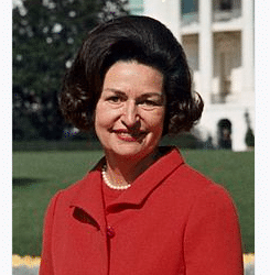 A woman in a red suit with dark hair smiles in front of a blurred background of a building and garden. The text reads Lady Bird Johnson and Official portrait, 1967.