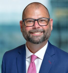 A bald man with a beard and glasses is smiling. He is wearing a blue suit, white shirt, and pink tie. The background is blurred, suggesting an indoor setting.