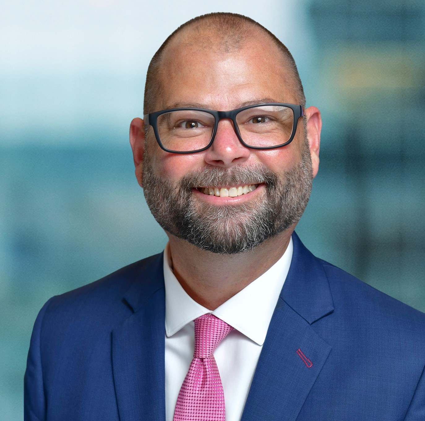 A bald man with a beard and glasses is smiling. He is wearing a blue suit, white shirt, and pink tie. The background is blurred, suggesting an indoor setting.