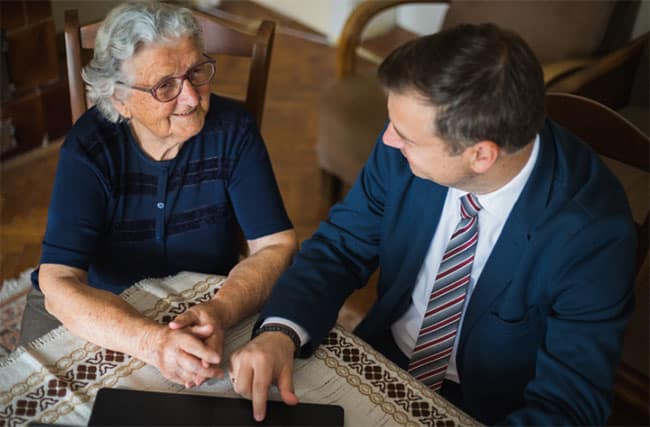An elderly woman and a man in a suit sit at a table with a patterned tablecloth. The man points at a digital tablet, engaging with the woman who is smiling. They appear to be having a friendly conversation indoors.