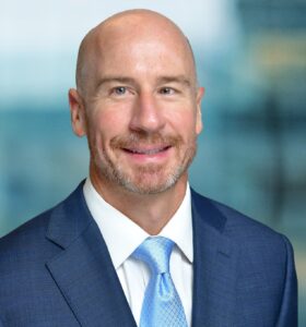 A bald man with a beard is smiling, dressed in a blue suit and light blue tie, standing against a blurred background.
