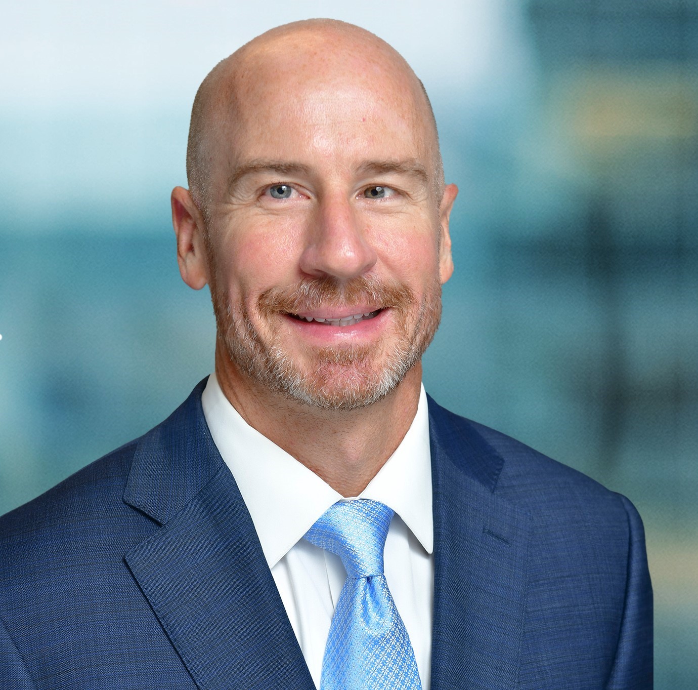 A bald man with a beard is smiling, dressed in a blue suit and light blue tie, standing against a blurred background.
