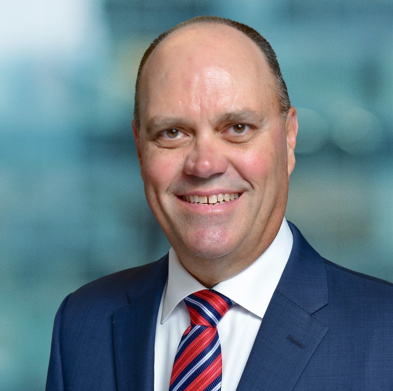 A smiling man in a blue suit and red striped tie stands in front of a blurred indoor background.